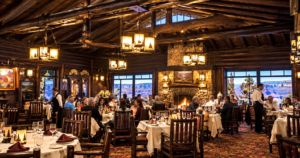 Rustic lodge restaurant interior featuring log beams, stone fireplace, warmly lit dining tables, guests enjoying meals, and expansive canyon views through large windows.