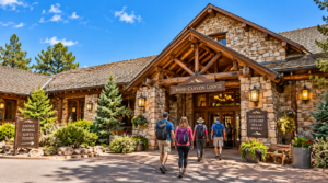 Grand Canyon Lodge entrance on a sunny day with hikers walking toward the rustic stone-and-timber building surrounded by pine trees, flowers, and clear blue skies.