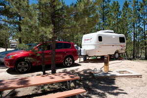 White Escape travel trailer parked beside a red SUV at a wooded RV campsite near Bryce Canyon, Utah, featuring tall pine trees, a picnic table, and an electrical hookup for comfortable camping.