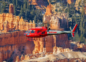 Red Zion Helicopters sightseeing helicopter flying past towering orange limestone hoodoos and rugged rock formations near Bryce Canyon National Park, Utah, with evergreen forests and dramatic canyon scenery in the background.