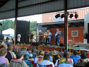 Beacon Rock WA. Camping And Hiking Tours 6 Bluegrass musicians performing on an outdoor covered stage with fiddles, guitars, and a banjo while festival visitors watch from folding chairs.