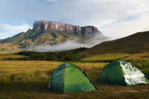 Two green camping tents set in an open field with Mount Roraima visible in the distance under cloudy skies.
