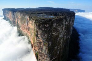 Mount Roraima viewed from above, surrounded by thick clouds with sheer cliffs rising from the rainforest and mist drifting across the plateau.