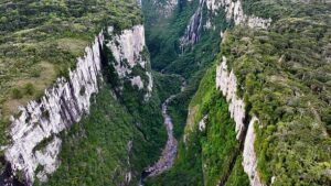 Scenic view of Itaimbezinho Valley in Brazil with dramatic canyon cliffs, a waterfall flowing from the canyon wall, rainforest vegetation, wildlife, and a campsite overlooking the valley during sunrise.