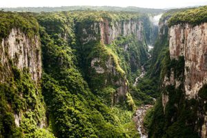 Scenic view of Itaimbezinho Valley in Brazil with dramatic canyon cliffs, a waterfall flowing from the canyon wall, rainforest vegetation, wildlife, and a campsite overlooking the valley during sunrise.