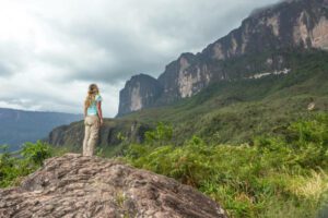 Woman stands on a large rock, looking up at Mount Roraima under cloudy skies, surrounded by rugged terrain.