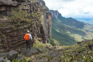 Person with a backpack stands on a rocky ledge, looking out over a lush valley with Mount Roraima rising in the distance under cloudy skies.