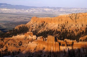 Panoramic view of Bryce Canyon National Park in Utah, featuring towering orange and golden rock formations, rugged canyon walls, evergreen trees, and distant mountain ranges beneath a pale blue sky.