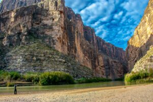 Person standing by a river at the base of a canyon with steep walls and surrounding vegetation.