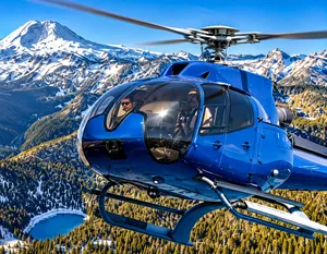 Blue helicopter flying above a dramatic snow-covered mountain range with alpine forests and a mountain lake below.