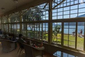 Guests relaxing inside a lakeside lodge while looking through large windows toward the lake, mountains, and people enjoying the waterfront.
