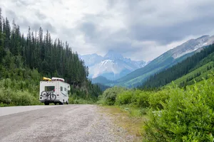 RV traveling along a scenic mountain road surrounded by dense evergreen forest, green valleys, and rugged snow-capped peaks.