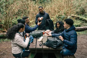 Group of hikers taking a break at a wooden picnic table in a forest, sharing food and organizing backpacks and hiking gear.