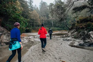 Three hikers walking along a sandy riverbank surrounded by dense evergreen forest, rocky terrain, and misty wilderness scenery.