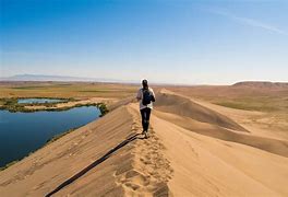 Bruneau Sand Dunes, Idaho, Camping & Hiking 3 Person walking with backpack in the desert with a water hole nearby on a partly cloudy day.