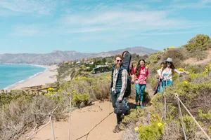 Kure Beach, NC, Camping And Hiking Tours 3 Group of hikers walking along a scenic coastal trail overlooking a sandy beach and blue ocean.