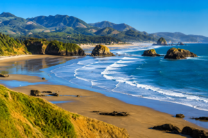 Scenic coastal landscape featuring a sandy beach, dramatic sea stacks, rolling ocean waves, rugged cliffs, and distant mountains under a clear blue sky.