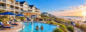 Corolla Beach, NC, Camping And Hiking Tours 8 Beautiful oceanfront resort with private balconies, a sparkling outdoor swimming pool, guests relaxing on lounge chairs, and visitors walking along a wooden boardwalk toward the sandy beach at sunset