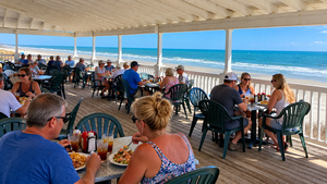 Visitors enjoying seafood and drinks at an oceanfront restaurant in Garden City Beach, South Carolina, with outdoor dining tables, a covered wooden deck, and beautiful views of the sandy beach and Atlantic Ocean.