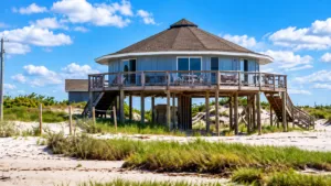 Cedar Island Beach NC, Camping & Hiking Tours 6 Elevated coastal beach cottage surrounded by sandy dunes and beach grass, featuring a wraparound deck and large windows beneath a bright blue sky.