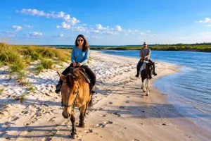 Cedar Island Beach NC, Camping & Hiking Tours 2 Two people horseback riding along a sandy coastal shoreline with calm blue water, grassy dunes, and clear skies.