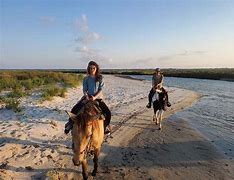 Two people riding horses on a beach next to the ocean with partly cloudy skies
