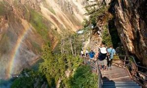 People walking downstairs on a side of a mountain with trees and a rainbow and steep cliffs.