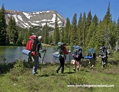 People with backpacks hiking near a river with trees nearby and a large mountain in the background.