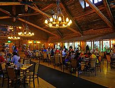 People sitting a restaurant in chairs eating at tables with large chandeliers for lights.