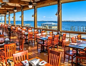 Bright waterfront restaurant interior with wooden tables and chairs overlooking blue water, docks, and a marina through large windows.
