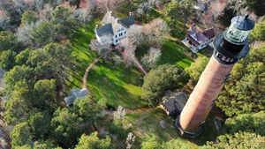 Lighthouse Inlet Beach, SC, Camping and Hiking Tours 7 Aerial view of Currituck Beach Lighthouse in Corolla, North Carolina, featuring its towering red-brick structure, historic buildings, winding pathways, and lush green grounds.