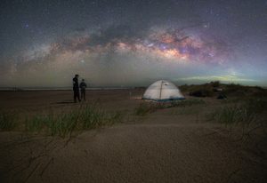 Fish Haul Beach SC Camping and Hiking Tours 6 Spectacular nighttime beach scene featuring a glowing camping tent on sandy dunes, two people stargazing, coastal grasses, and the colorful Milky Way stretching across a star-filled sky.