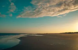 Pea Island Beach, NC, Camping & Hiking Tours 1 Pea Island Beach, North Carolina, at sunset with a wide sandy shoreline, gentle Atlantic Ocean waves, reflective tidal water, coastal dunes and a dramatic golden sky.