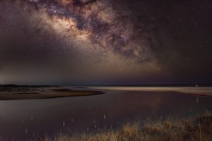 Corolla Beach, NC, Camping And Hiking Tours 7 Spectacular Milky Way stretching across a star-filled night sky above a peaceful coastal beach, with calm water reflecting starlight and sandy dunes covered in beach grass.