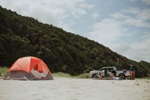 Corolla Beach, NC, Camping And Hiking Tours 2 Orange and gray camping tent pitched on a sandy beach near wooded dunes, with a pickup truck and campers enjoying an outdoor adventure.