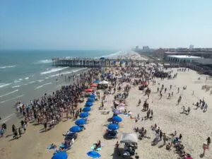 Figure Eight Island Beach, NC, Camping & Hiking Tours 5 Helicopter tour view from above showing a busy sandy beach, ocean waves, beachfront activity, and a long pier extending into the water.