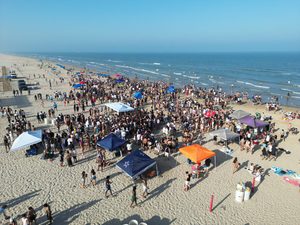 Corolla Beach, NC, Camping And Hiking Tours 5 Aerial view of a large crowd gathered on a sandy ocean beach, with colorful canopy tents, beach umbrellas, parked vehicles, and people enjoying the shoreline.
