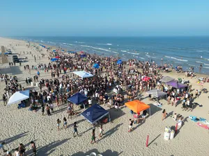 Large crowd gathered on a wide sandy beach with colorful canopy tents, ocean waves, and people enjoying activities along the shoreline.