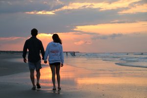 Isle of Palms, South Carolina: Beach Camping Getaways, Coastal Adventures & Scenic Escapes 4 A couple holding hands while walking barefoot along a sandy ocean beach at sunset, with gentle waves, colorful orange and pink clouds, and golden reflections shimmering across the wet shoreline.