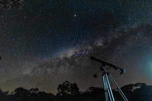 Stargazing telescope positioned beneath a spectacular star-filled night sky, with the Milky Way stretching across the darkness above silhouetted trees and a peaceful natural landscape.