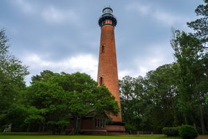 Lighthouse Inlet Beach, SC, Camping and Hiking Tours 3 Tall historic red-brick lighthouse surrounded by lush green trees and grassy grounds beneath a cloudy sky.