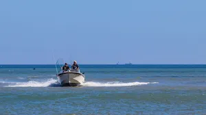 Two anglers riding in a small fishing boat across calm blue water under a clear sky, with fishing rods and equipment onboard.