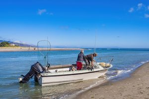 Fish Haul Beach SC Camping and Hiking Tours 2 Two fishermen preparing their small motorboat along a sandy shoreline, with fishing rods, a large landing net, calm blue coastal waters, and a clear sunny sky.