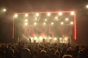 Large outdoor concert crowd watching a live band perform on a brightly illuminated stage with dramatic red and golden lights.