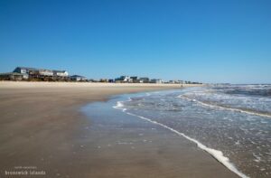 Sandy beach with gentle ocean waves and buildings visible in the distance.