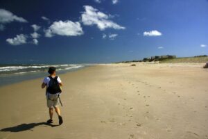Person with a backpack walking along a sandy beach beside the ocean.
