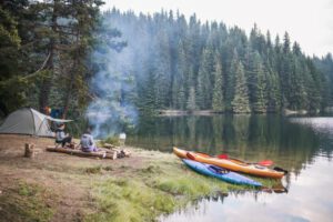 people camping next to a lake with canoes and a fire burning and a tent in the woods
