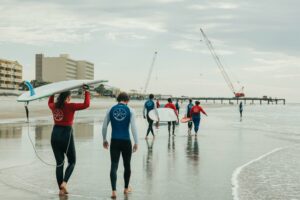 people walking on the beach with beach boards next to the ocean with buildings and a peer