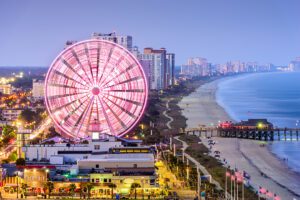 Ferris wheel and coastal buildings at a sandy beach with a pier extending into the ocean.