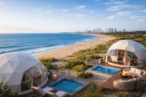 Two white geodesic glamping domes on wooden decks overlooking a sandy beach, with coastal dunes, Atlantic waves, and Punta Del Este skyline in the background.